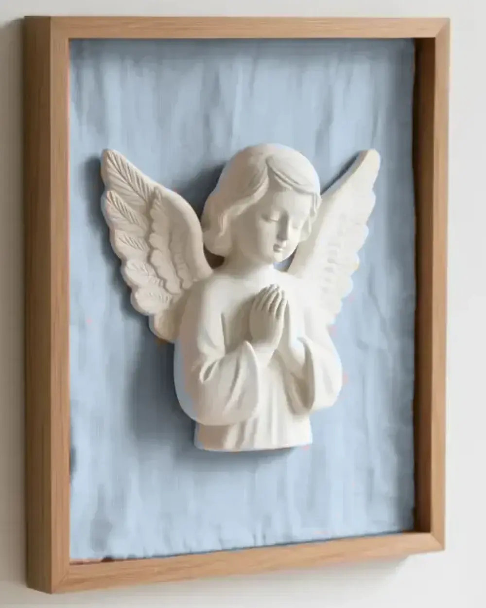 Primer plano del relieve del Ángel de la Guarda en escayola blanca, con las manos juntas, sobre tela azul novena dentro de un marco de madera clara