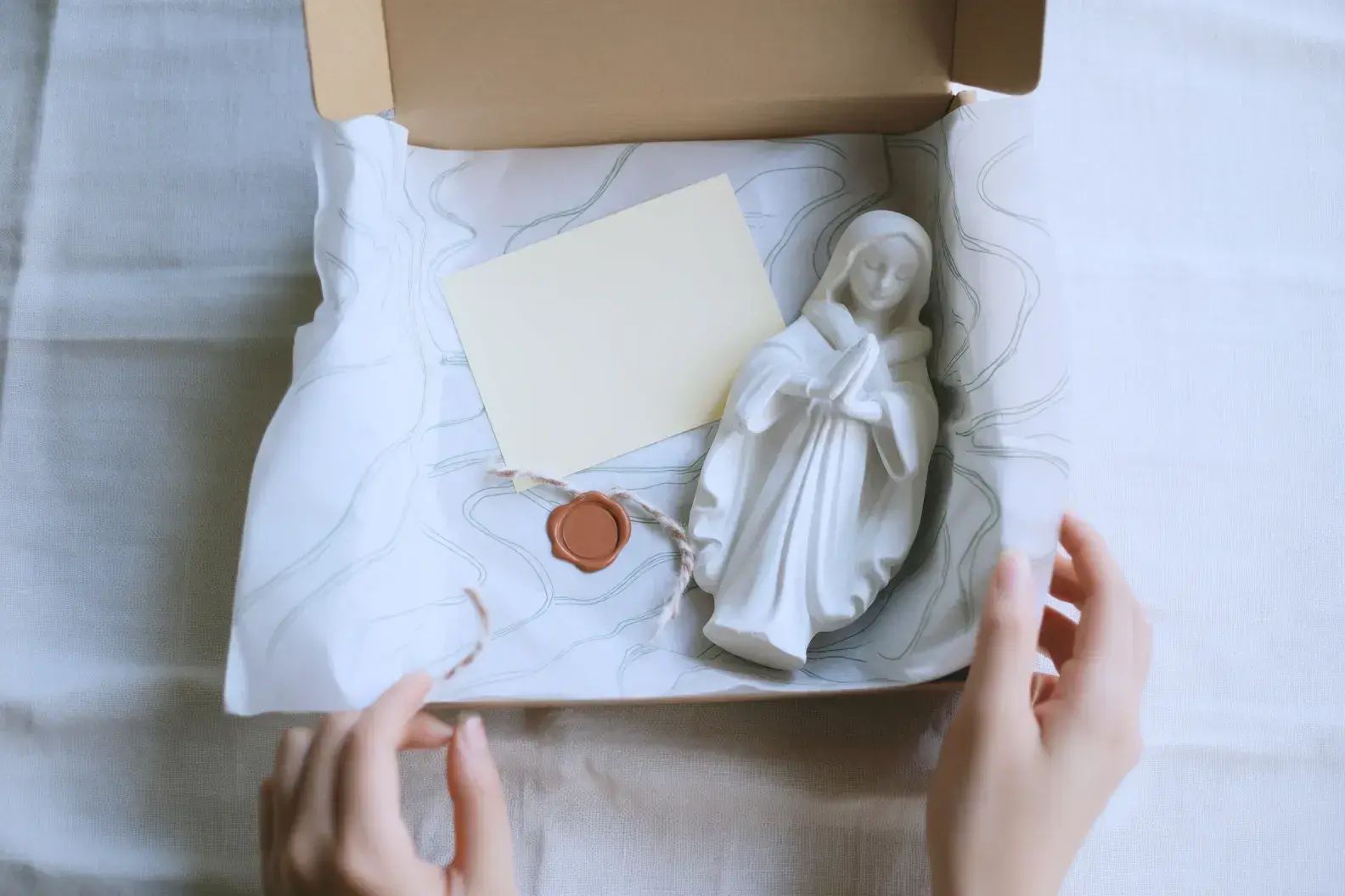 Manos abriendo una caja de cartón con papel de seda estampado, una tarjeta y una figura de escayola blanca dentro
